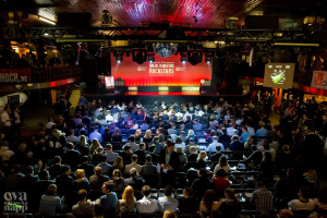 Eine große Menschenmenge, die auf Stühlen vor einer Bühne mit Bildschirmen, Bannern und Geländern sitzt, die von Lichtern beleuchtet wird und gespannt auf den Beginn eines Online-Marketing-Rockstars-Events wartet, mit einem Wasserzeichen in der linken unteren Ecke.