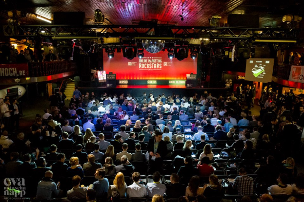 Eine große Menschenmenge, die auf Stühlen vor einer Bühne mit Bildschirmen, Bannern und Geländern sitzt, die von Lichtern beleuchtet wird und gespannt auf den Beginn eines Online-Marketing-Rockstars-Events wartet, mit einem Wasserzeichen in der linken unteren Ecke.