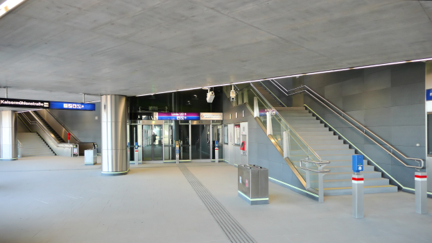 Eine belebte Straße in der Stadt mit starkem Verkehr, ein Zug auf Höhgleisen, umgeben von Bäumen, Gebäuden, Baustellenabsperrungen, Strommästen und Bahninfrastruktur.