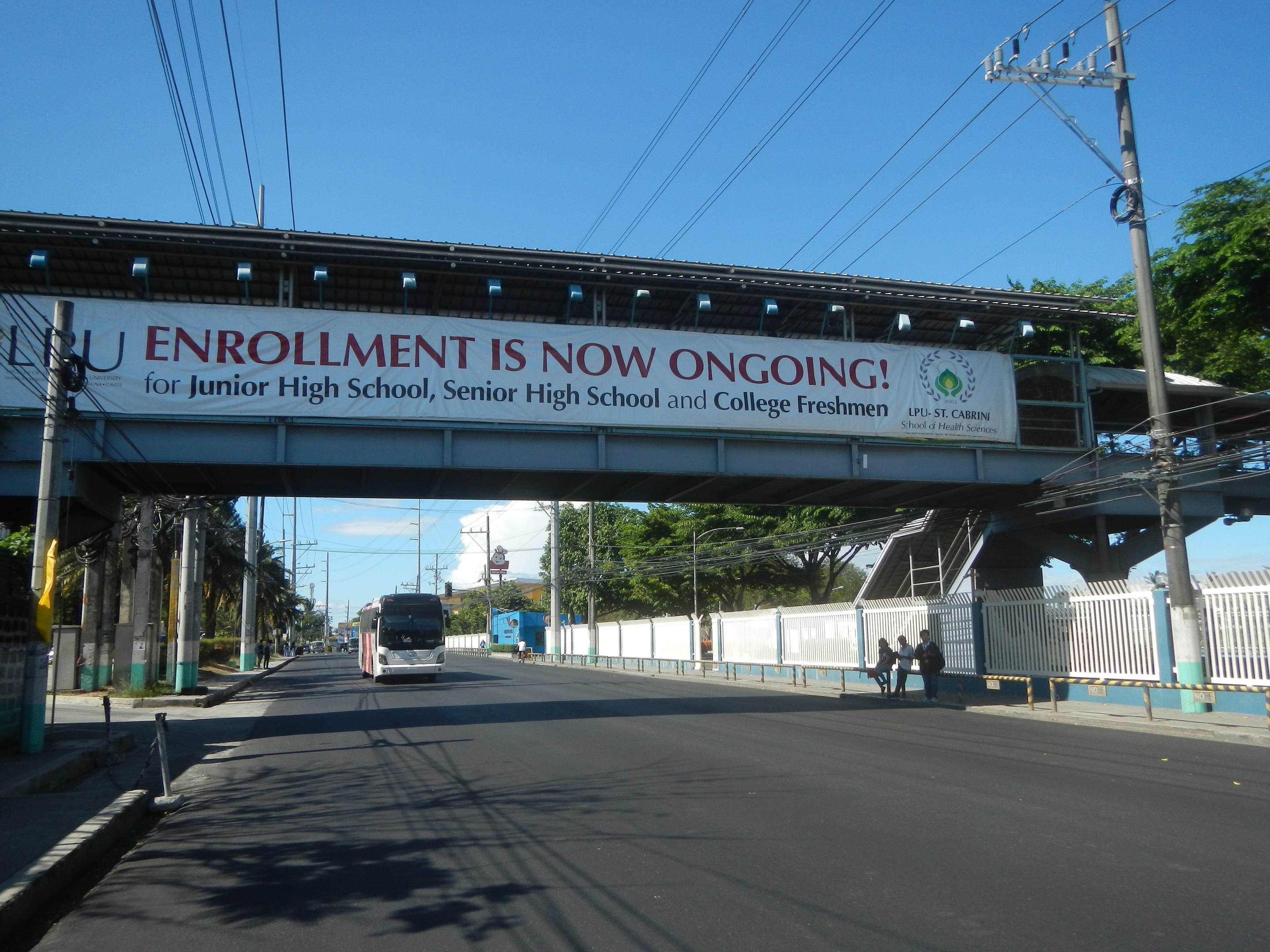 Ein Bus fährt unter einer Brücke mit einem "Jetzt ist die Anmeldung möglich"-Schild, Menschen gehen auf dem Gehweg darunter, Strommasten mit Drähten, Bäume und ein klarer blauer Himmel im Hintergrund.