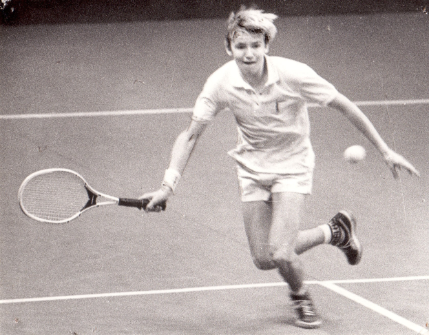 Ein junger Mann in einem weißen Tennis-Outfit schlägt mit einem Schläger einen Ball in der Luft, mit einem entschlossenen Gesichtsausdruck.