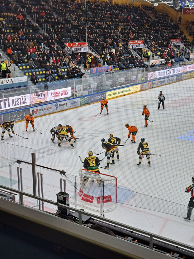 Eine Gruppe von Menschen spielt Hockey auf einer Indoor-Eisfläche mit einem Netz, umgeben von einem Zaun, unter Stadionbeleuchtung, mit Zuschauern im Hintergrund und Schildern.