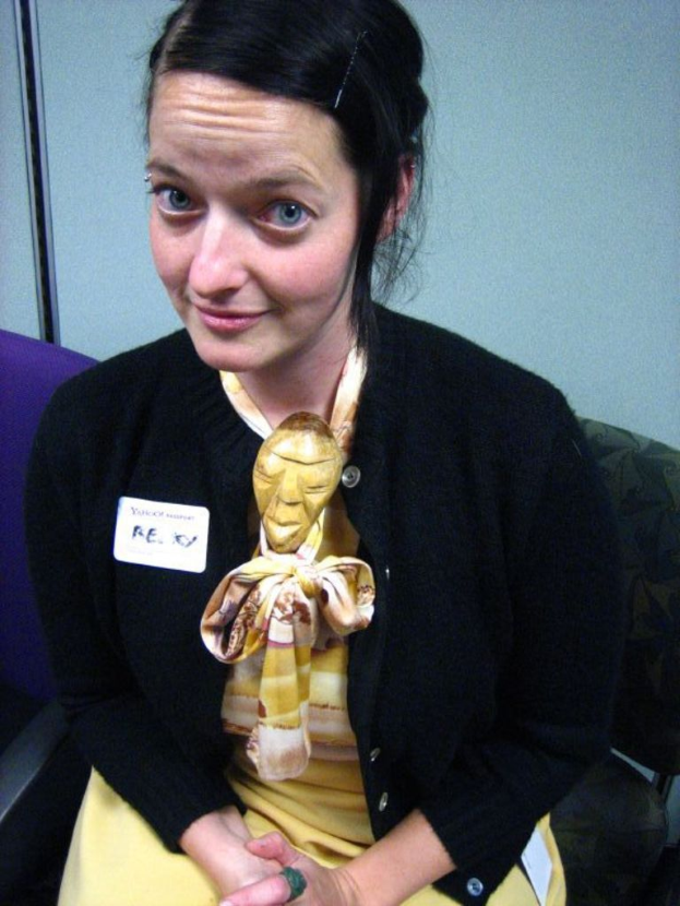 Eine Frau sitzt auf einem Stuhl und trägt eine schwarze Jacke und ein gelbes Kleid, mit einem Anstecker an ihrer Kleidung, vor einer Wand.