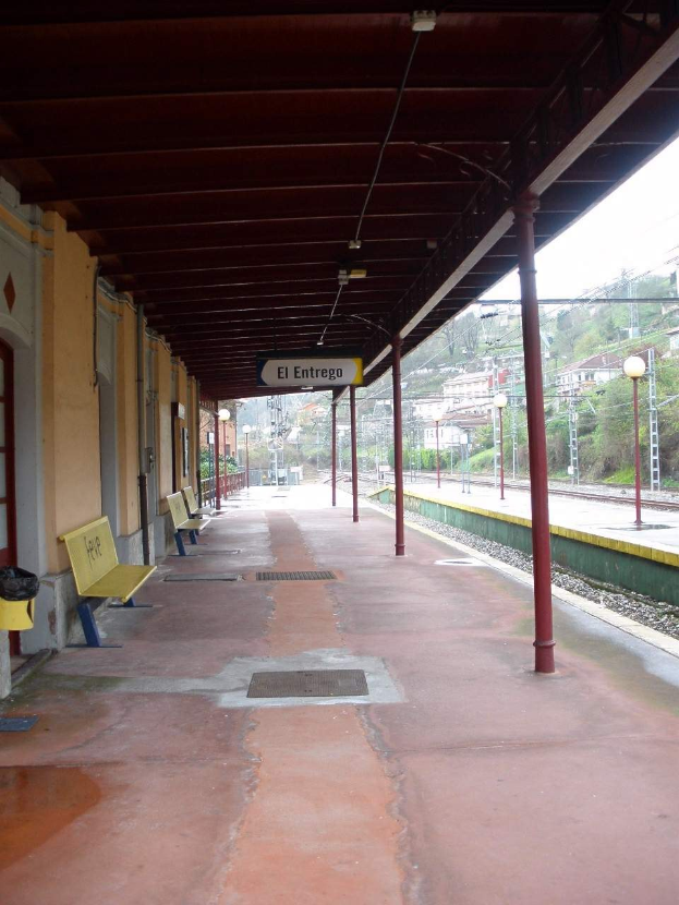 Ein Bahnsteig mit Bänken, einem Schild, Pfosten, Lampen und einer Anzeigetafel, umgeben von einem Gebäude mit Türen und Fenstern, vor Bäumen, anderen Gebäuden und dem Himmel.