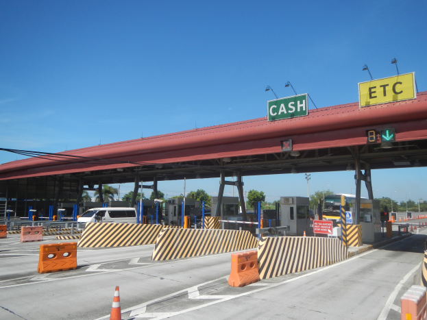 Eine Mautstation mit einem "Barzahlung"-Schild, umgeben von Fahrzeugen, Schrankenstangen, Verkehrszeichen, Straßenlaternen, Strommasten, Stromkabeln, Bäumen und einem bewölkten Himmel.
