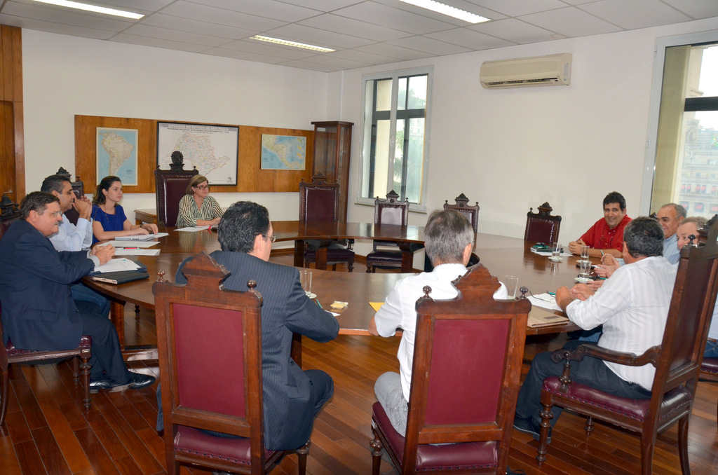 Eine Gruppe von Menschen, die an einem Tisch sitzen und Papiere und Gl√§ser vor sich haben, ein Klimager√§t, eine Anschlagtafel und Karten an der Wand im Hintergrund.