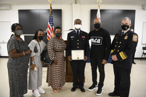 Gruppe von maskierten Menschen vor Flaggen, mit einer Person in der Mitte, die ein Zertifikat hält.