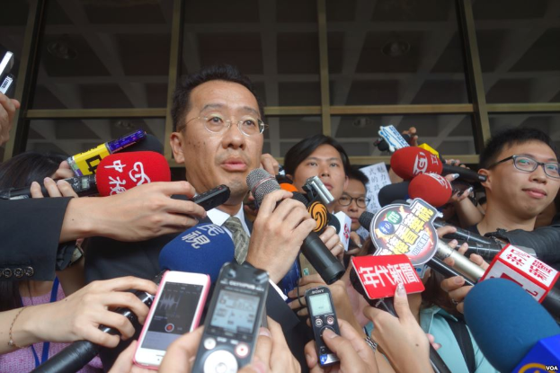 Ein Mann im Anzug und Krawatte mit Brille steht umgeben von Reportern, einige halten Mikrofone und Mobiltelefone, mit einer Glaswand im Hintergrund.