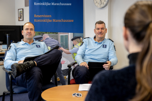 Zwei Polizeibeamte sitzen an einem Tisch mit Papieren und Gegenständen und sprechen mit einer Frau, mit einer Wandtafel, Uhr und Fotorahmen im Hintergrund und einem weiteren Tisch mit Monitor, Tastatur und Maus links daneben.