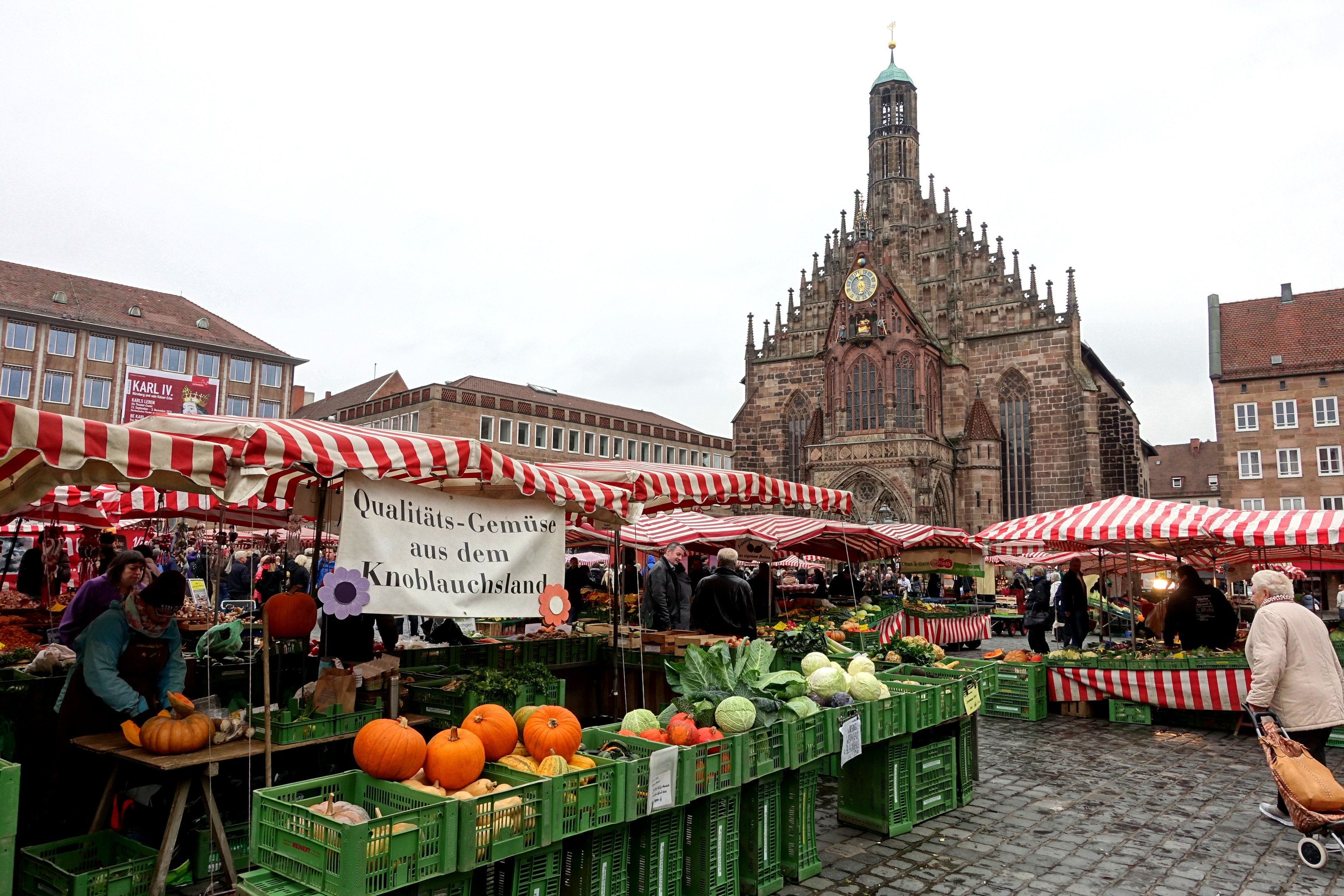 Ein belebter Markt in Nürnberg, Deutschland, mit Ständen, die verschiedene Obst- und Gemüsesorten anbieten, Menschen mit Taschen und aufgestellte Zelte im Hintergrund, mit Gebäuden und einem Uhrenturm.