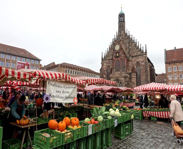 Ein belebter Markt in Nürnberg, Deutschland, mit Ständen, die verschiedene Obst- und Gemüsesorten anbieten, Menschen mit Taschen und aufgestellte Zelte im Hintergrund, mit Gebäuden und einem Uhrenturm.