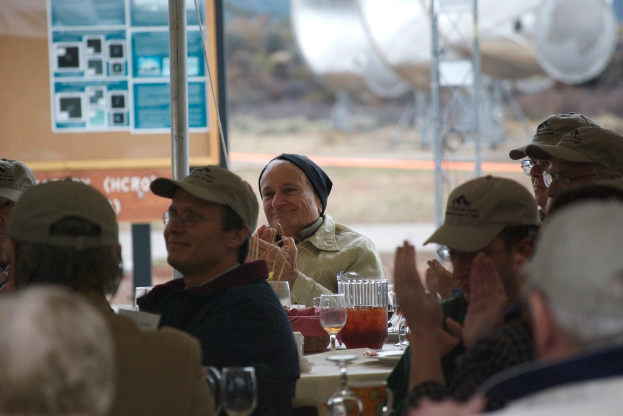 Menschen, die auf Stühlen sitzen und klatschen, mit einem Tisch daneben, der Biergläser, einen Teller und ein Tuch enthält, sowie einem Pfahl, einer Platte und Gras im Hintergrund.