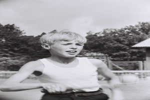 Ein junger Junge in Schwarz und Weiß spielt Tennis auf einem von Bäumen und einer Hütte umgebenen Platz, hält einen Schläger mit einem Ball in der Luft.