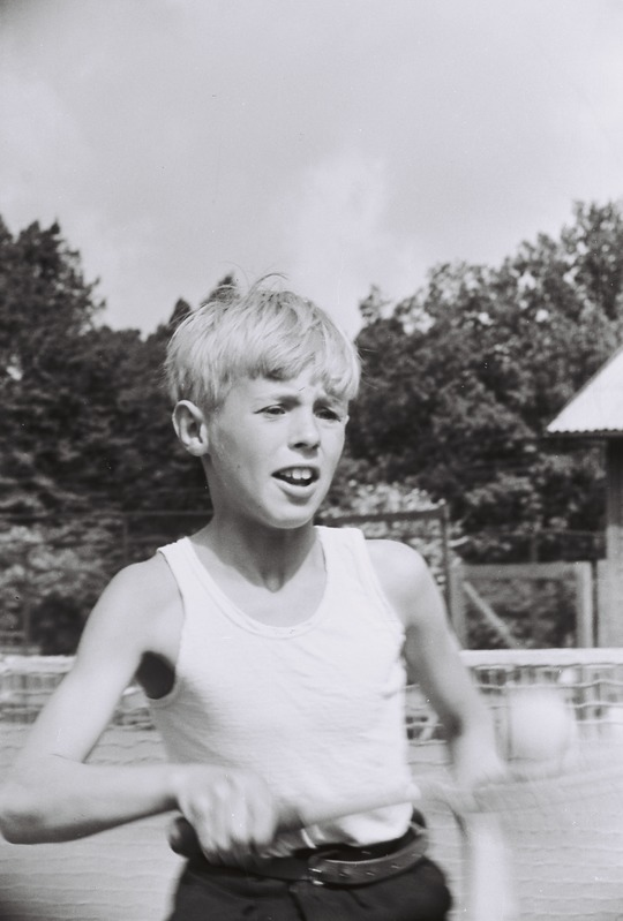 Ein junger Junge in Schwarz und Weiß spielt Tennis auf einem von Bäumen und einer Hütte umgebenen Platz, hält einen Schläger mit einem Ball in der Luft.