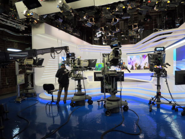 Ein Mann steht vor einem Fernseher und hält ein Mikrofon in der Hand, umgeben von Kameras auf dem Boden, einem Stuhl, einem Bildschirm, einer Wand und einer Decke mit Lampen, was auf eine Live-Übertragung hindeutet.