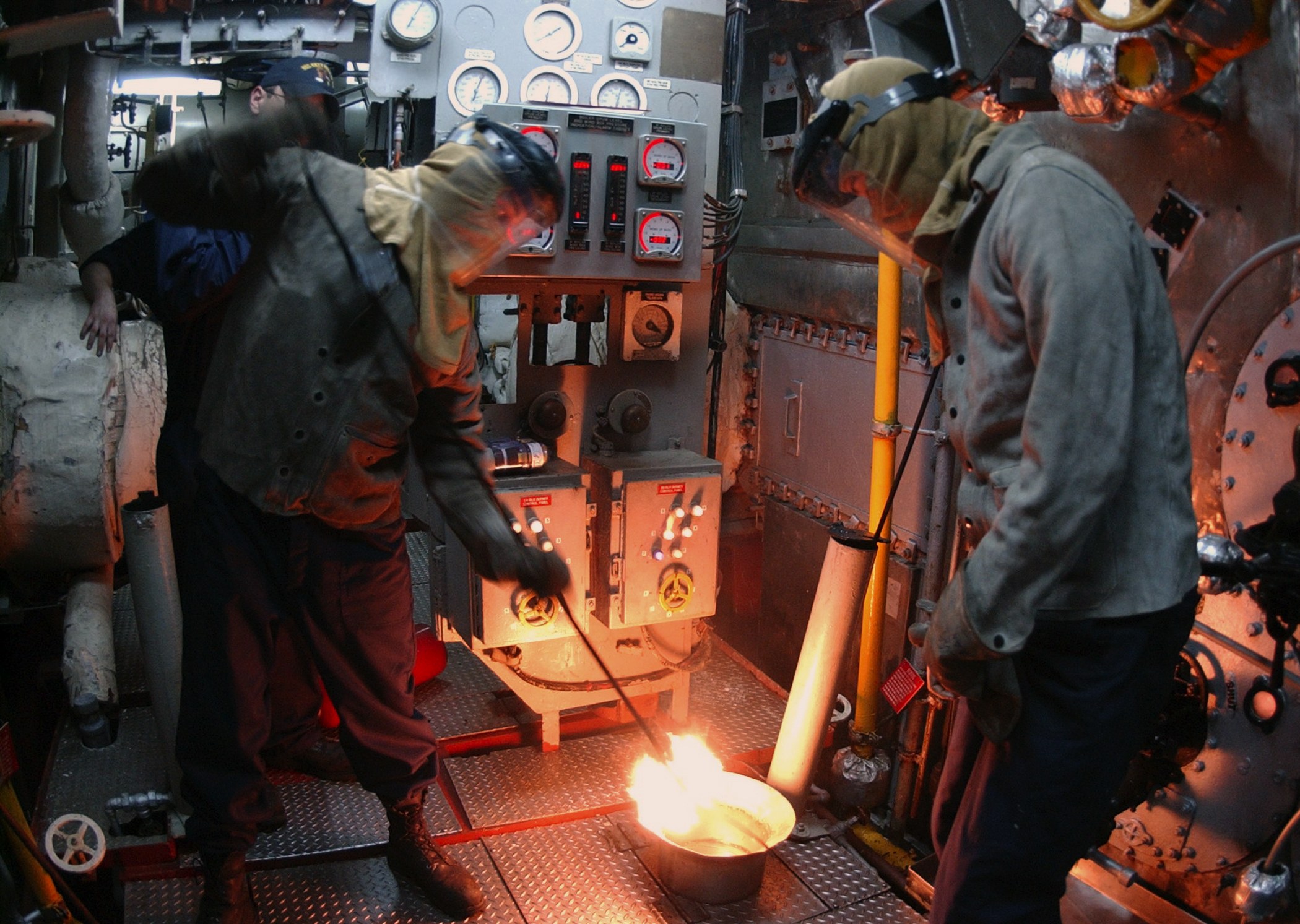 Zwei Männer in Schutzausrüstung arbeiten an einer Maschine in einer Fabrik mit einem Feuer vor ihnen und Messgeräten im Hintergrund.