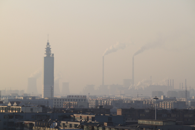 Luftaufnahme einer Stadt mit Gebäuden und Bäumen im Vordergrund, Rauch aus Schornsteinen gegen einen nebligen grauen Himmel.