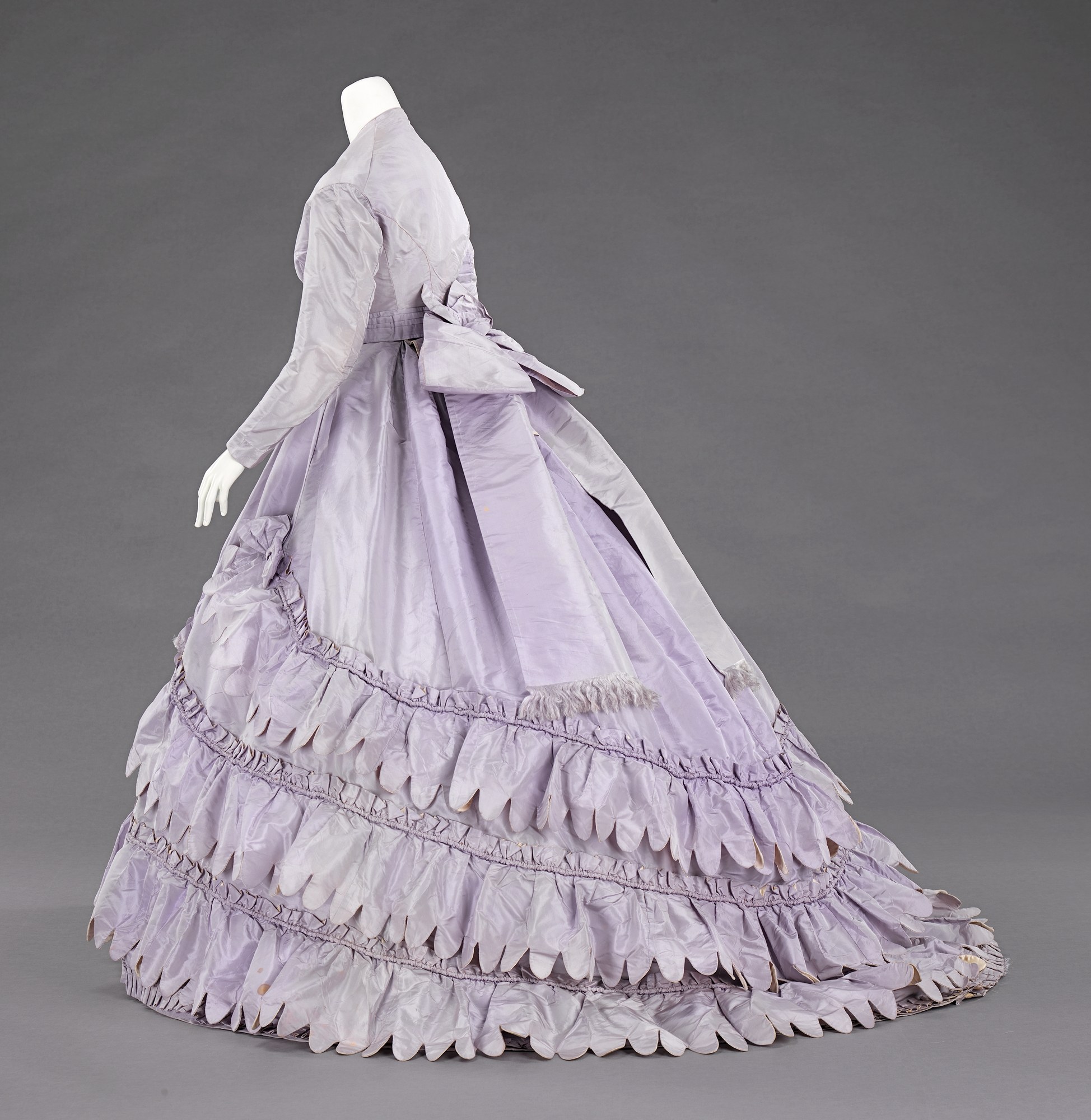Eine Schaufensterpuppe in einem lila, langärmligen Ballkleid mit engem Oberteil, vollem gerüschtem Rock und leichtem Stoff vor einem grauen Hintergrund.