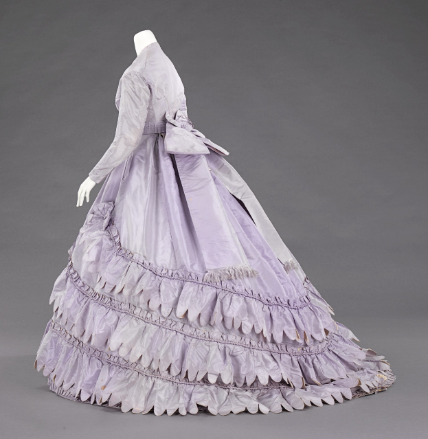 Eine Schaufensterpuppe in einem lila, langärmligen Ballkleid mit engem Oberteil, vollem gerüschtem Rock und leichtem Stoff vor einem grauen Hintergrund.