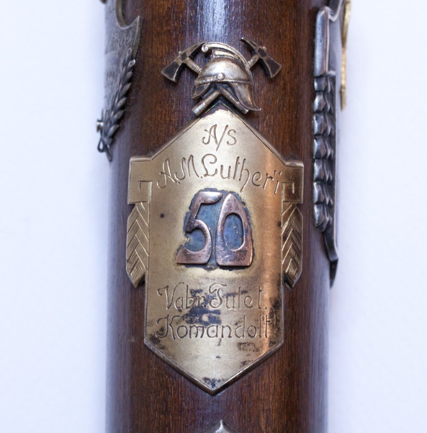 Nahaufnahme eines braunen Gewehrs mit einer Metallplakette, auf der "50. Jahrestag der deutschen Armee" steht, vor einem weißen Hintergrund.