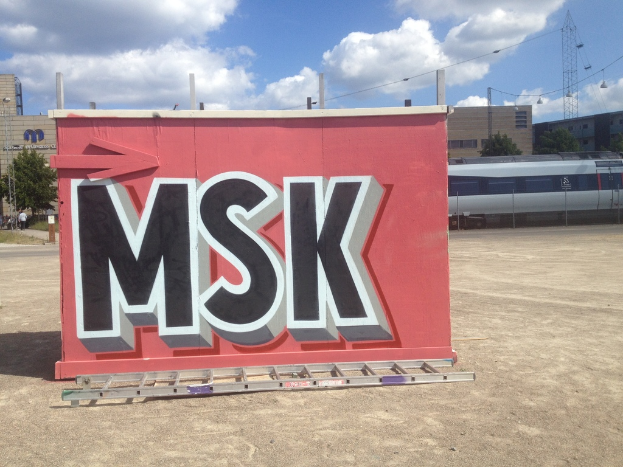 Ein Schild in der Mitte mit einer Leiter davor, Gebäude und Bäume im Hintergrund, ein Turm rechts und ein Zug in der Ferne.