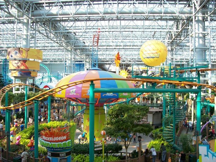 Großes Indoor-Wasserpark mit einer Achterbahn, umgeben von Menschen, Bäumen, Pflanzen und beleuchteten Deckenstangen.
