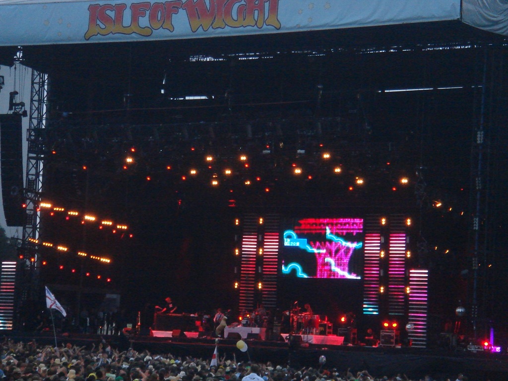 Eine Bühne mit Scheinwerfern, einem Bildschirm und Musikern, die Instrumente spielen, mit einem Banner oben, einer Menge unten und Fahnen mit Stangen und Ballons drumherum.