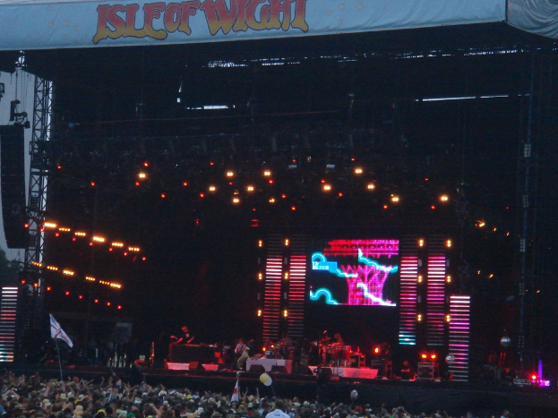 Eine Bühne mit Scheinwerfern, einem Bildschirm und Musikern, die Instrumente spielen, mit einem Banner oben, einer Menge unten und Fahnen mit Stangen und Ballons drumherum.