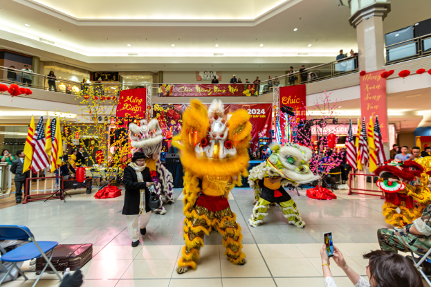 Ein lebendiger Chinese New Year-Feier in einem Einkaufszentrum mit tanzenden Menschen in Kostümen und anderen, die mit Mobiltelefonen sitzen, umgeben von Flaggen, Bannern, Dekorationen und Lichtern.