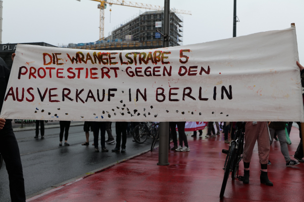 Gruppe von Menschen mit einem Banner "Die Wrangellstraße 5 Protestiert Gegen den Ausverkauf in Berlin" auf einer Straße mit Fahrrädern, Pfählen, Gebäuden und einem Kran im Hintergrund.