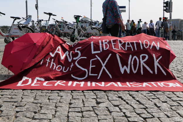 Gruppe von Menschen protestiert mit Regenschirmen, Fahrrädern und einer roten Fahne mit der Aufschrift "Befreiung ohne Entkriminalisierung der Sexarbeit" auf einer Stadtstraße.