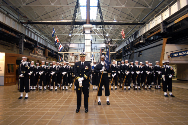 Eine Gruppe uniformierter Männer mit Mützen und Gewehren steht in Formation in einem großen Raum mit Flaggen, Texttafeln an den Wänden und Deckenleuchten und Stangen.
