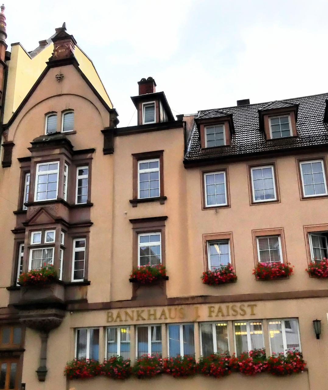 Ein mehrstöckiges Gebäude mit zahlreichen Fenstern und Blumen an der Seite, das sich in Frankfurt, Deutschland befindet und ein Schild trägt, auf dem "Bankhaus I Faist" steht, sowie einen sichtbaren Himmel oben.