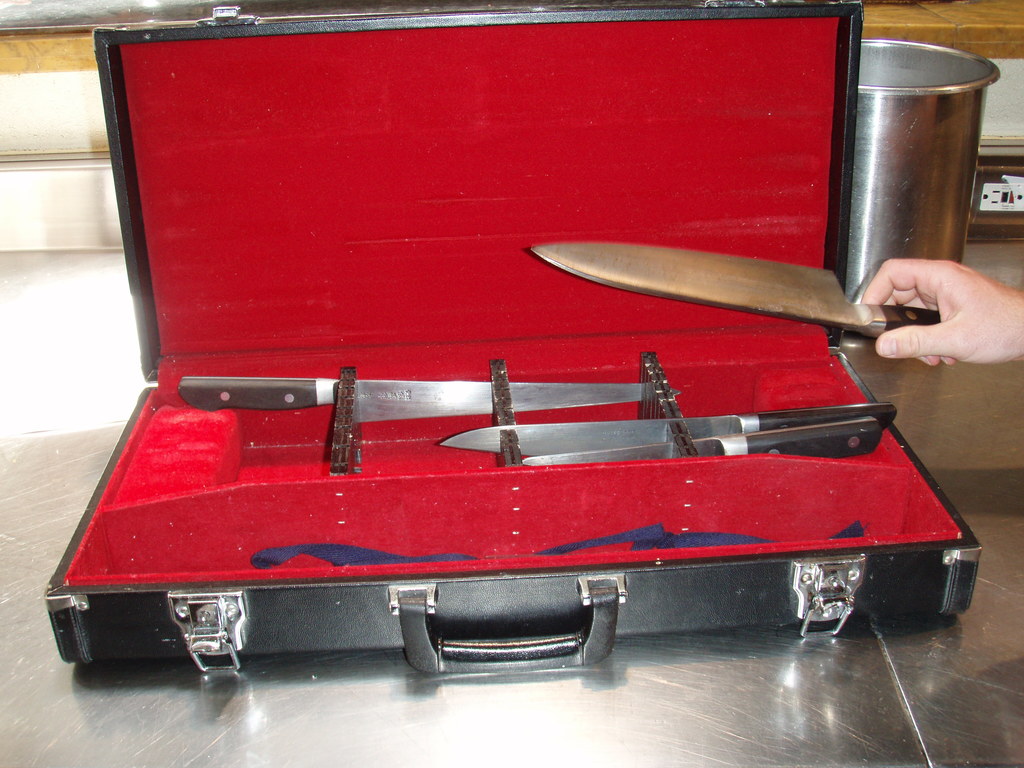 Ein Koffer mit einem Messer und rotem Tuch oben drauf, ein Glas und eine Hand im Hintergrund.