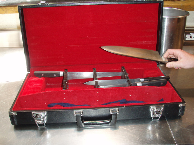 Ein Koffer mit einem Messer und rotem Tuch oben drauf, ein Glas und eine Hand im Hintergrund.