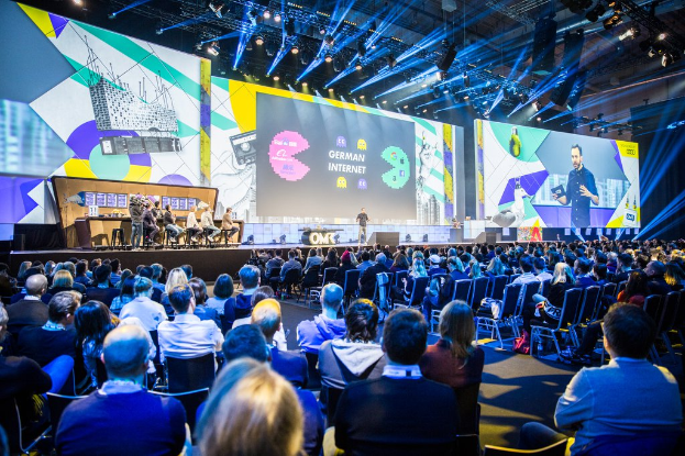 Eine große Menschenmenge sitzt auf Stühlen vor einer Bühne, einige Personen stehen auf dem Podium, ein Tisch mit Gegenständen links und Bildschirme und Lichter im Hintergrund auf der Deutschen Internet-Konferenz 2019.