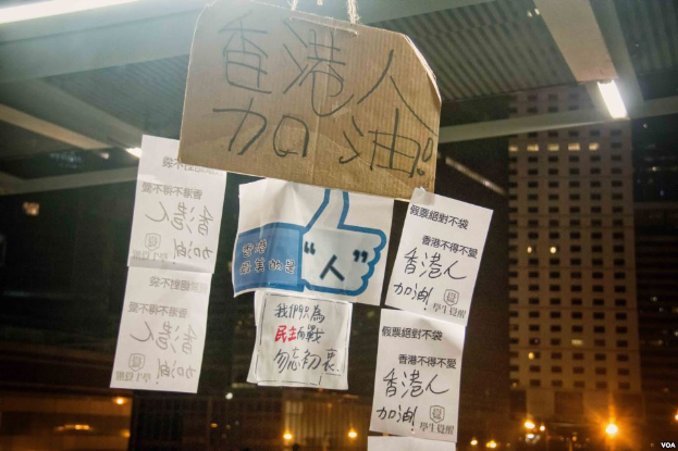 Eine Gruppe von Menschen steht vor einem Gebäude bei Nacht, beleuchtet von Stadtlichtern, mit mehreren Protestschildern an der Decke.
