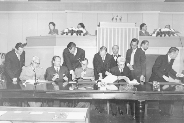 Eine Gruppe von Menschen, die auf Stühlen sitzen und andere hinter ihnen an einem Tisch mit Mikrofonen, Gläsern und Blumensträußen stehen.