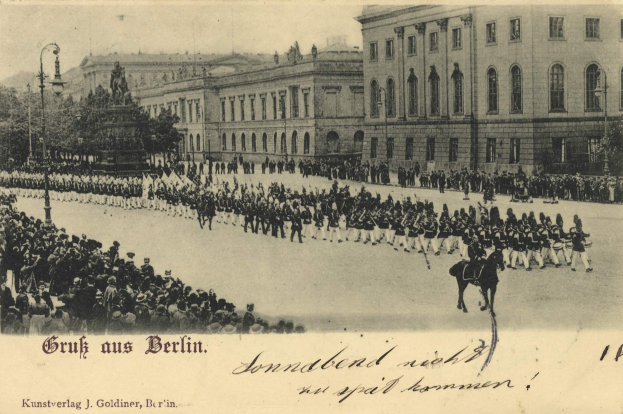 Ein altes Schwarz-Weiß-Foto von einer Parade in Berlin, Deutschland, zeigt Menschen auf dem Boden und auf Pferden, eine Statue auf einem Sockel, Straßenlaternen, Bäume, Gebäude mit Fenstern und einen bewölkten Himmel, mit Text am unteren Bildrand.