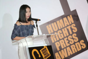 Eine Frau in einem schwarzen Kleid steht an einem Podium mit einem Mikrofon und hält eine Rede vor einem Banner "Human Rights Press Awards", ihr Haar ist zurückgebunden und ihr Gesichtsausdruck ernst und konzentriert.