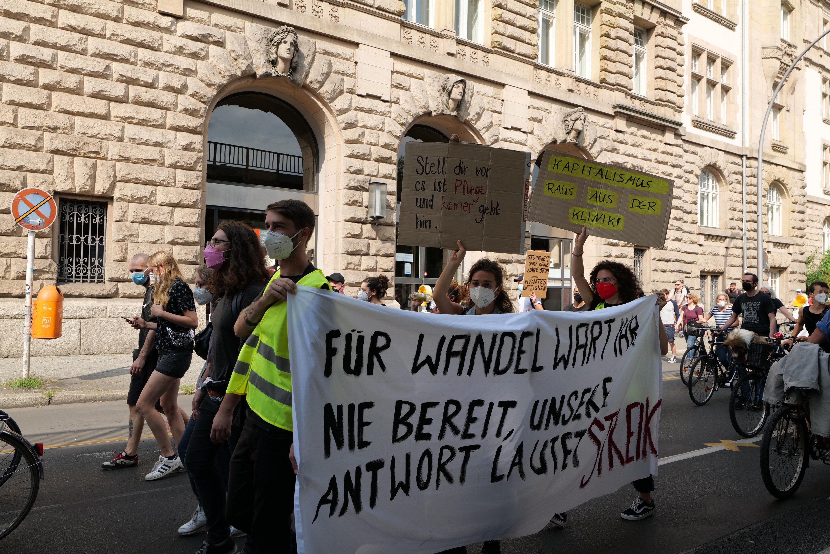 Eine Gruppe von Menschen marschiert bei einer Demonstration durch eine Straße, einige halten Schilder und andere fahren Fahrräder, im Hintergrund ein Gebäude mit Rundbogenfenstern, Säulen und Skulpturen.