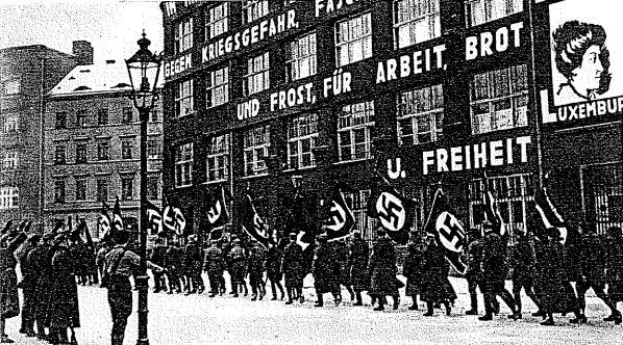 Schwarzes und wei├čes Bild einer Gruppe, die mit Fahnen und Transparenten die Stra├če entlangmarschiert, mit einem Lichtmast links und einer Tafel rechts mit der Aufschrift "Deutsche Nationalsozialistische Arbeiterpartei in Berlin".