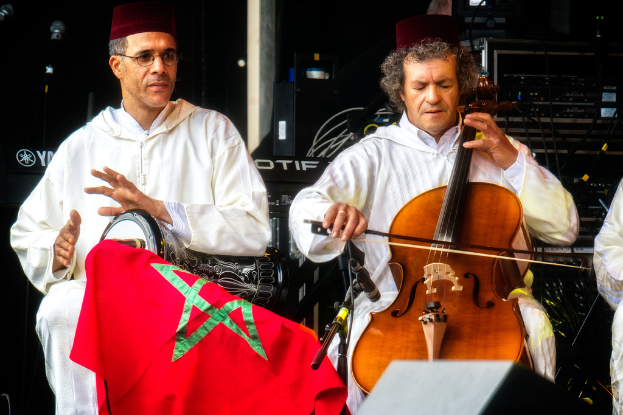 Zwei Männer in identischen Kostümen und Mützen sitzen nebeneinander und spielen verschiedene Musikinstrumente, hinter ihnen sind Musikgeräte zu sehen.