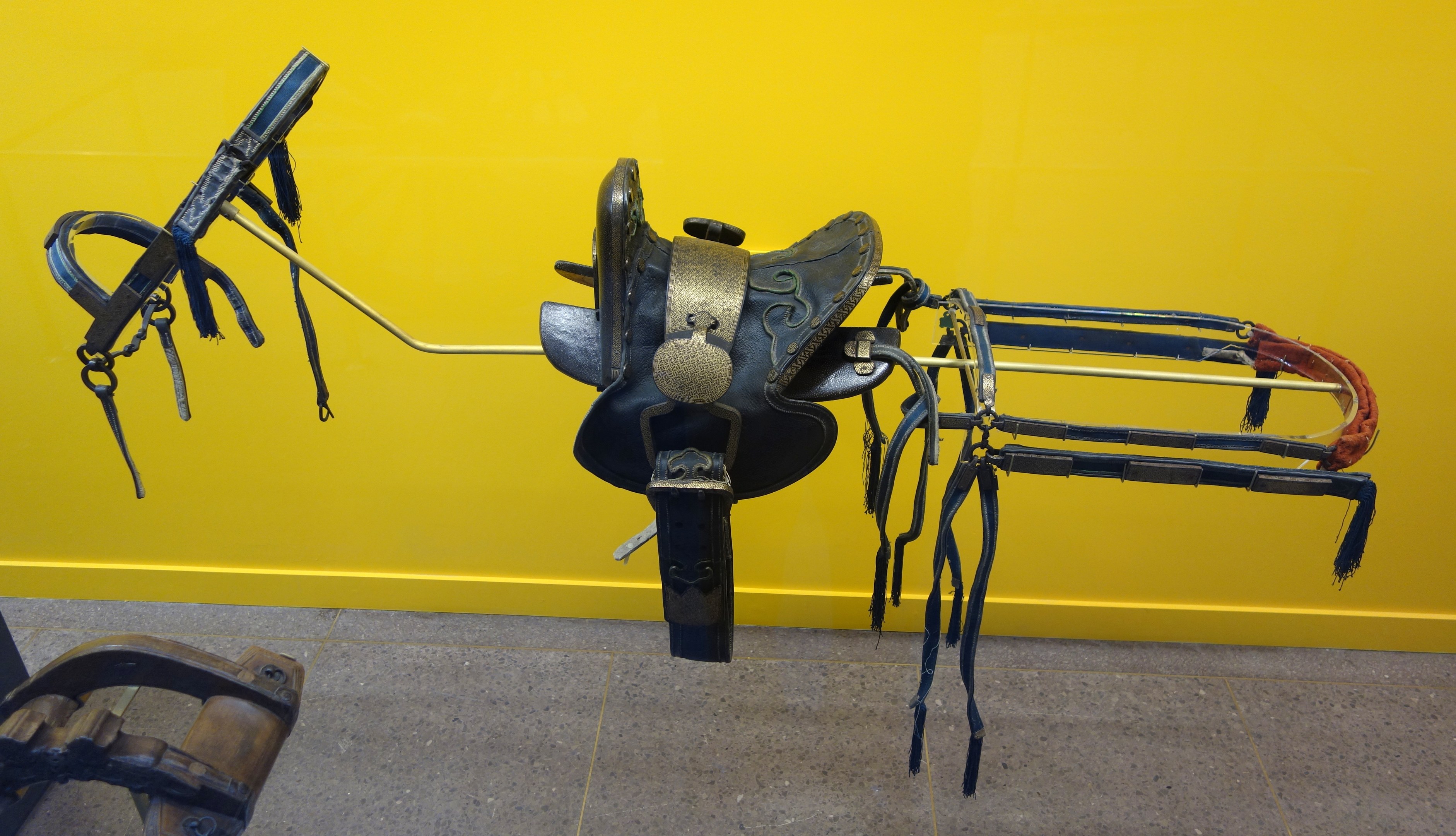 Pferdesattel in einem Museum ausgestellt mit einem Metallobjekt links und einer gelben Wand im Hintergrund, Boden unten sichtbar.