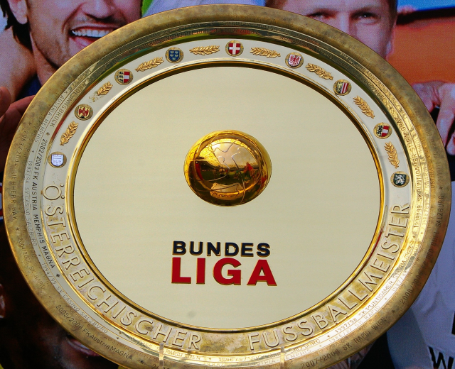 Goldener Schild mit "Bundesliga"-Text in den Händen eines Mannes, im Hintergrund ein Banner mit drei Personen.