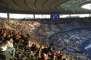 Eine große Menschenmenge sitzt im Allianz Stadion in München, Deutschland, und schaut ein Fußballspiel zu, mit einer Bühne auf der rechten Seite und Fahnen, Stangen und einem Bildschirm im Hintergrund.