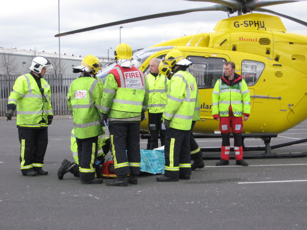 Eine Gruppe von Arbeitern in grünen Jacken steht neben einem Hubschrauber auf dem Boden, mit einem Zaun, kleinen Bäumen und einer Hütte im Hintergrund.