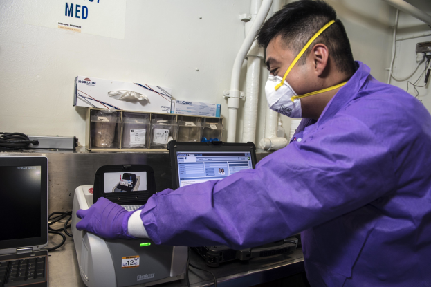 Ein Mann in einem violetten Laborkittel und -maske arbeitet an einem Laptop, umgeben von Laborgeräten, Kabeln und einer mit der University of California, Berkeley, beschrifteten Wand.