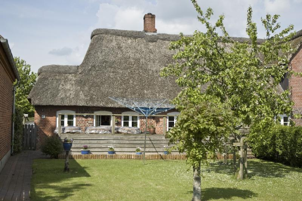Ein Reetdachhaus mit einem Garten davor, umgeben von saftigem grünen Gras, Bäumen und Pflanzen, mit Stufen, die zur Eingangstür führen, und Töpfen mit Pflanzen drumherum, unter einem Himmel voller weißer, flauschiger Wolken.
