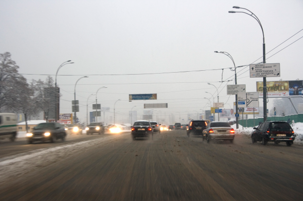 Eine städtische Straße voller Verkehr an einem verschneiten Tag, mit Fahrzeugen, schneebedeckter Straße, Laternen, Texttafeln, Bäumen, Gebäuden und einem Himmel im Hintergrund.